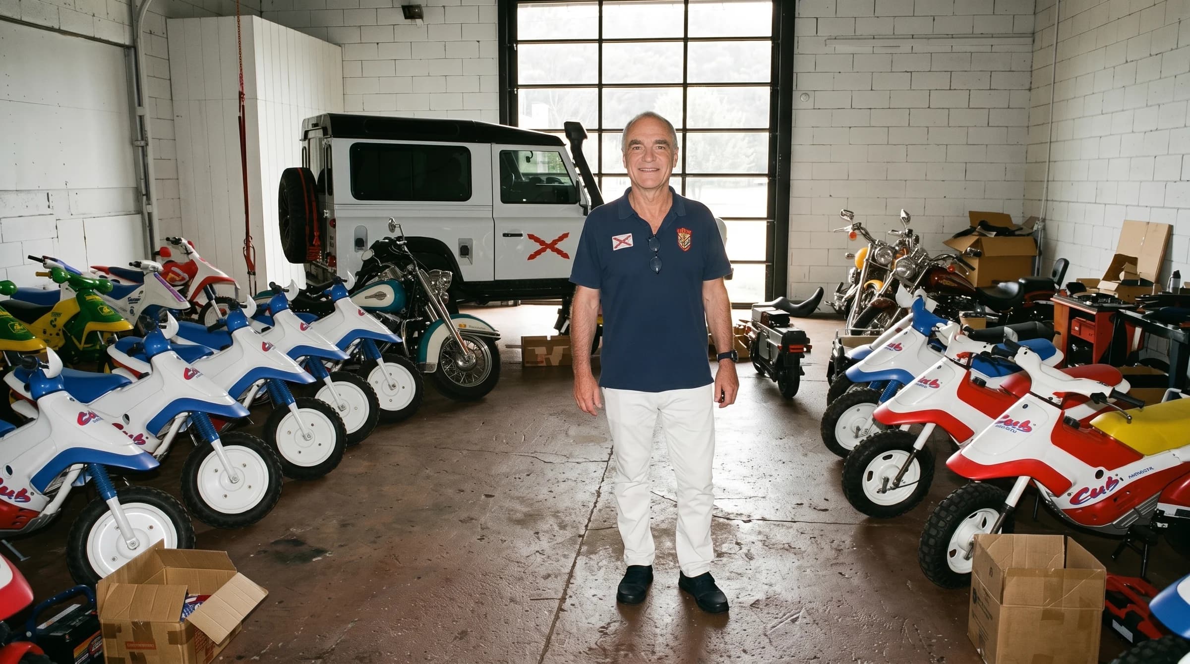 David Meunier standing among his collection of classic Honda Cub mini bikes in his warehouse, his white Land Rover Defender behind him, daylight through the industrial windows.
